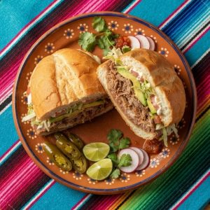 A torta on a telera roll cut in half, filled with shredded pork, beans, avocado, lettuce and tomato, with pickled jalapeños.